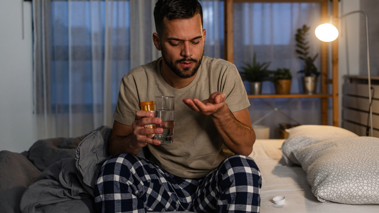 A man holding a pill and glass of water in his hands while sitting at the edge of his bed