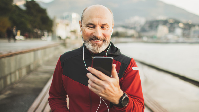 A healthy older man standing on a dock wearing headphones and looking at his cellphone