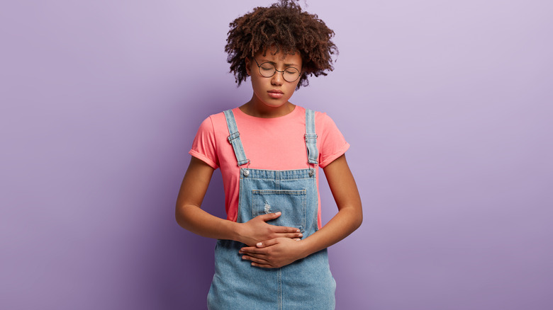 Woman stands with her hands to her abdomen in pain, against a purple background