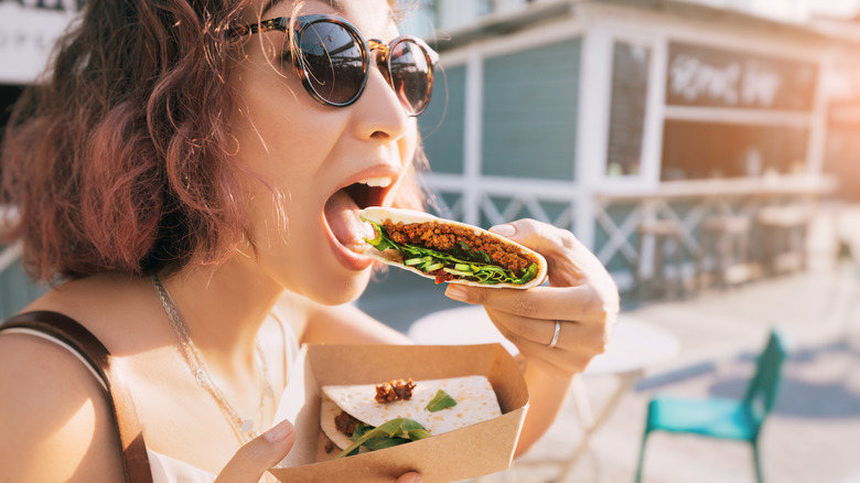 a young woman in sunglasses opens her mouth to bite into a taco