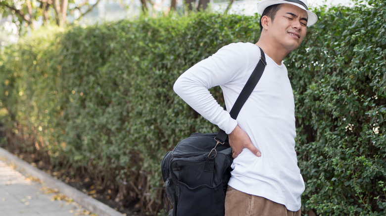 a man holds his back in pain as he carrys a big cross-shoulder bag 