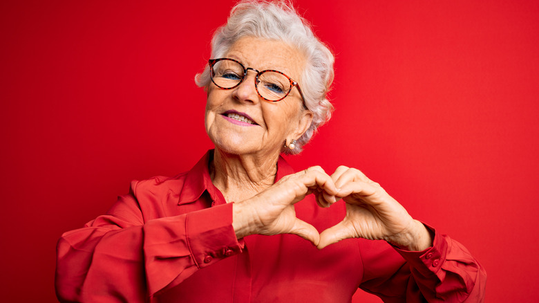 Older woman against red background smiling and making heart shape with hands
