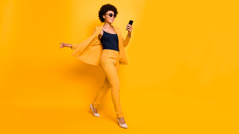 Fashionable woman wearing sunglasses, a suit, and stilettos smiling at her phone