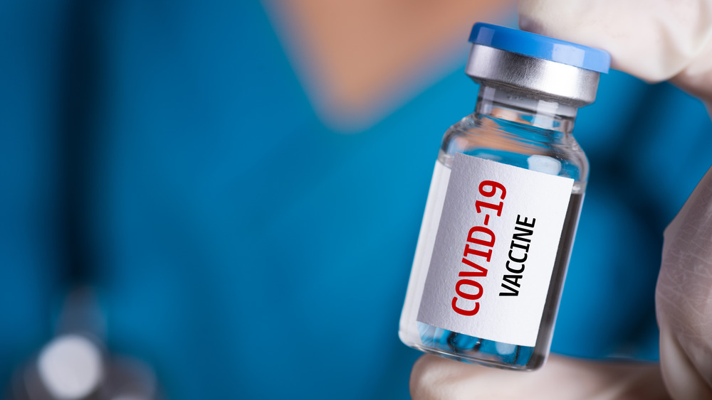 Gloved hand holding vial with "COVID-19 vaccine" written on it