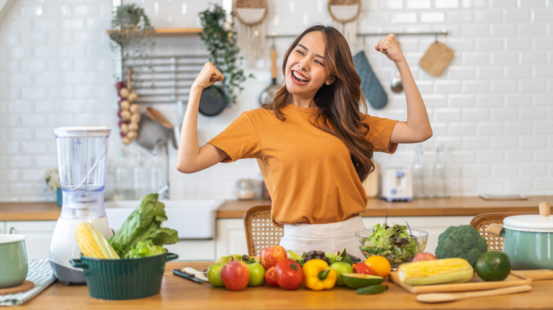 A healthy woman holding her arms up in strength while standing behind a table of fruits and vegetables