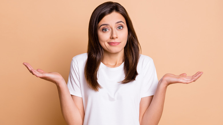 Woman shrugging against a beige backdrop looking directly at camera