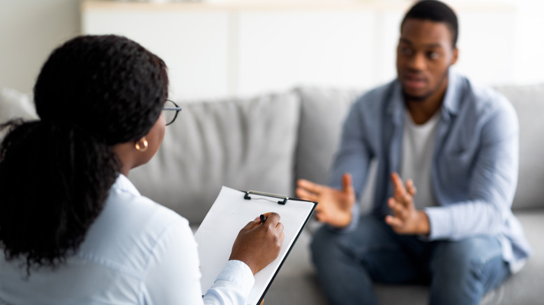 Young man sitting on a couch talking to a mental health provider holding a clipboard