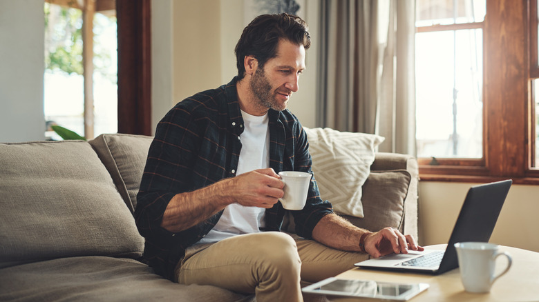 A man holding a teacup while working on his laptop and sitting on his couch
