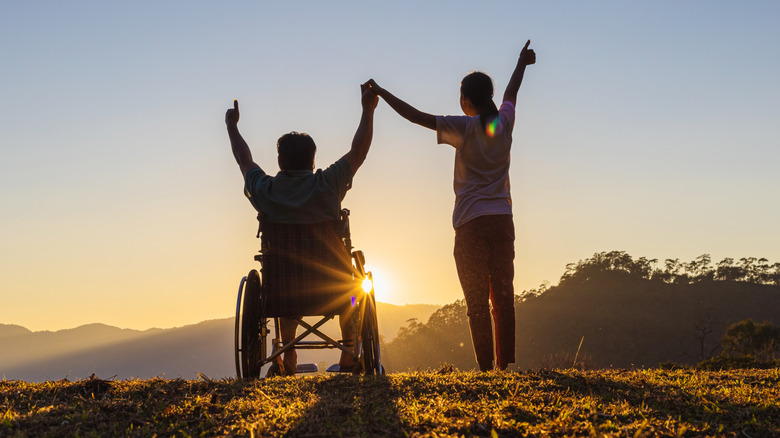 man in wheelchair and woman holding hands up triumphantly against a sunset