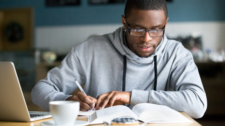 Man with glasses studying textbook near open laptop.