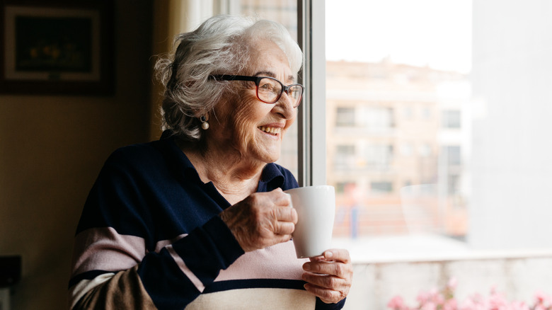 elderly woman holding a cup of tea and smiling while looking out a window
