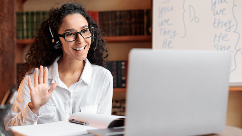 Woman seated at a desk wearing glasses and a headset waving and smiling at computer screen