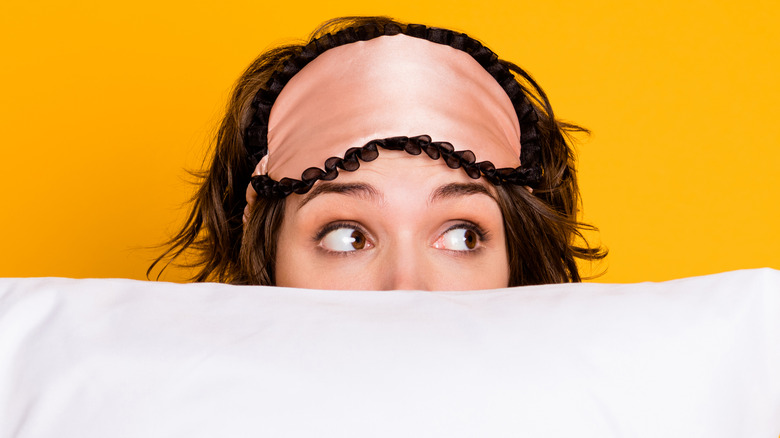 Close up on woman's eyes looking to the left with the rest of her face behind a pillow and wearing a sleep mask on her head against a yellow background