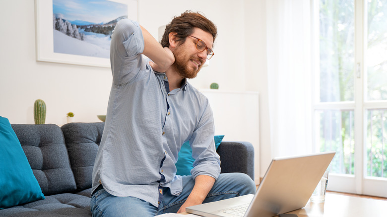 Man wearing glasses, sitting on couch holding his neck, laptop on coffee table