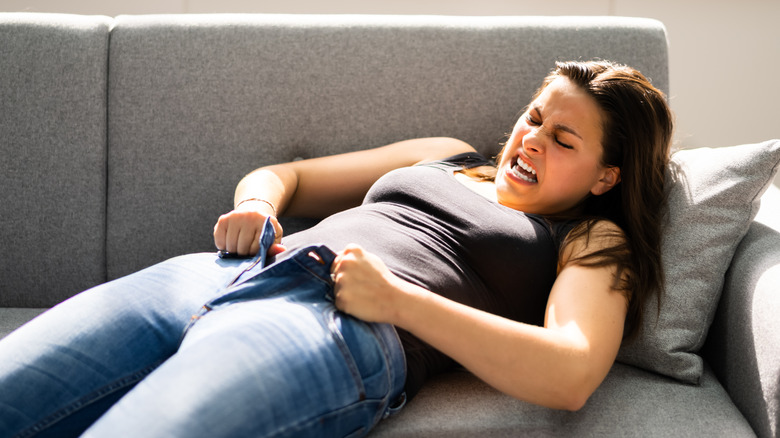 a woman grimaces while trying to put on pants that are too tight 