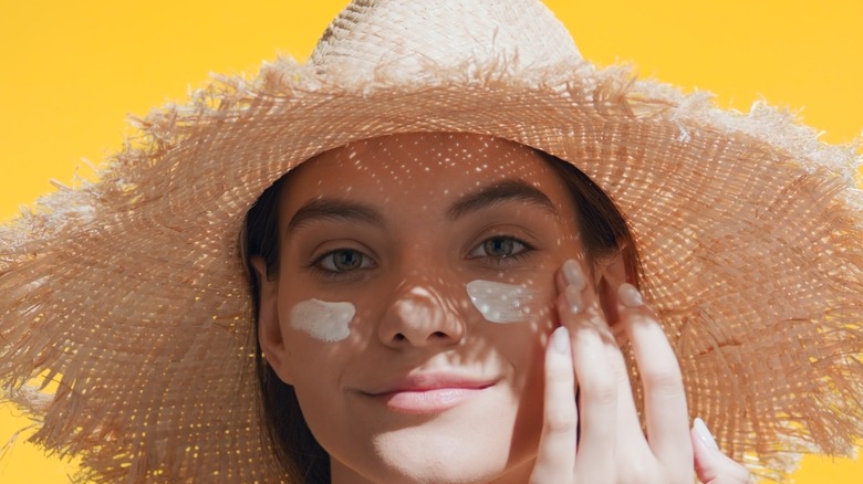 a woman in a straw hat applies sunscreen to face 