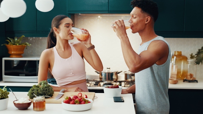 A man and woman drinking juice in the kitchen with fruits and vegetables on the counter