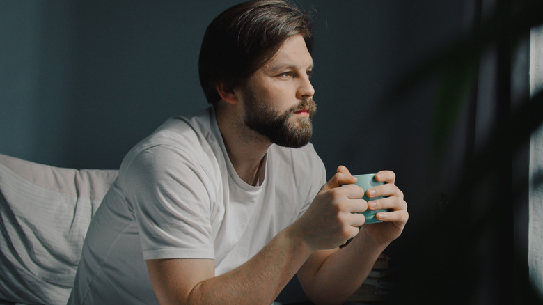 male adult sitting on edge of bed enjoying drink in mug