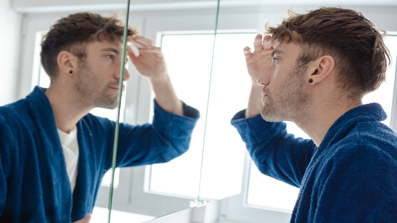 A concerned man in a blue robe looking in a mirror
