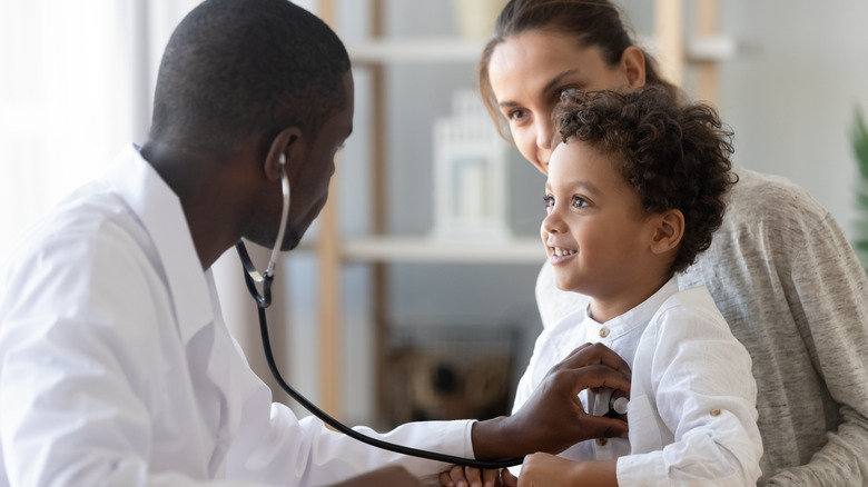 Pediatrician with stethoscope listens to lungs and heart of a young child sitting on their mother's lap