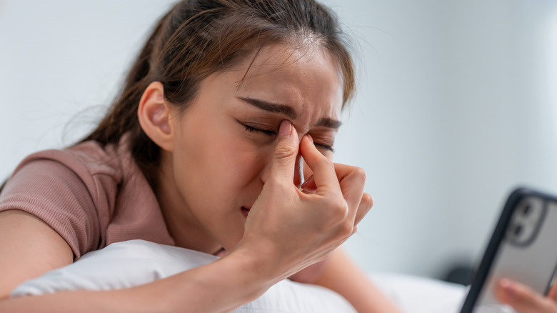 A woman lying in bed looking at her phone and rubbing her eyes