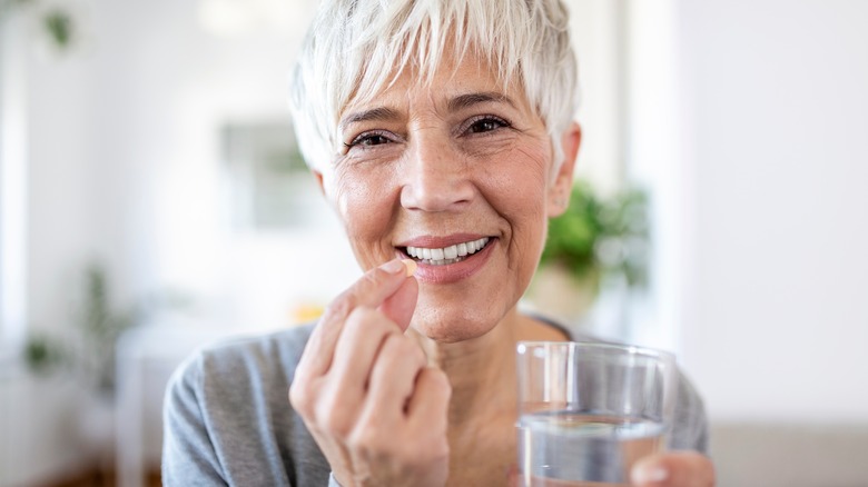 Older woman with short white hair smiles holding a pill and glass of water
