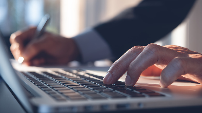 Hands typing on a laptop keyboard with pen in hand