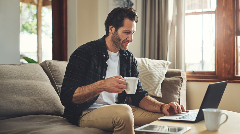 A man holding a teacup while working on his laptop while sitting on the couch