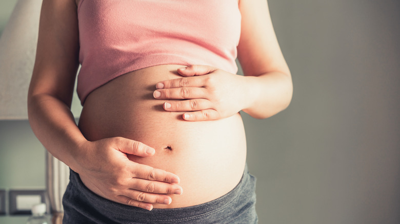 Pregnant person wearing a pink top and grey bottoms holding bare belly with both hands