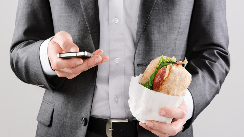 Business man working on the go with smart phone and take away sandwich lunch