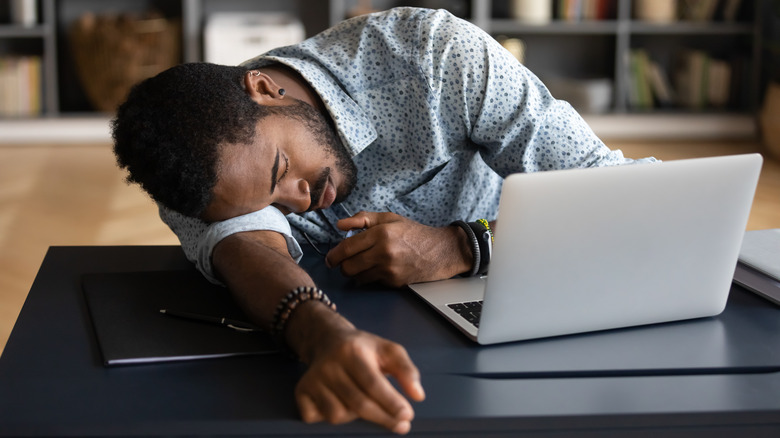 Young man asleep at his desk resting on his arm in front of his laptop