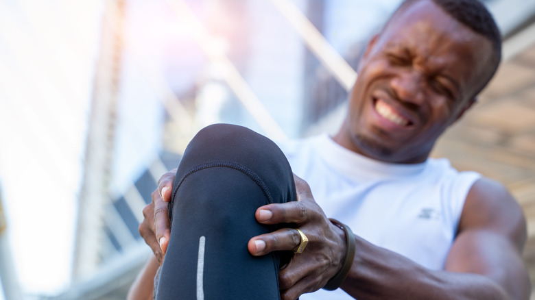Person wearing white sleeveless top and blue athletic pants is grimacing in pain and holding one knee