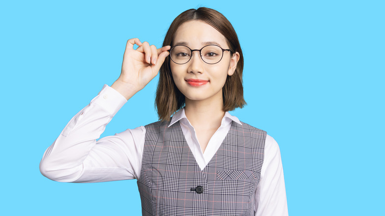 Person with short brown hair and a vest and long sleeved white shirt, holding onto their black eyeglasses with two fingers while standing in front of a blue background