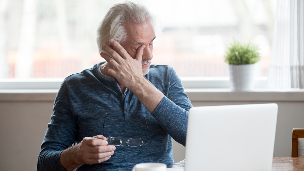 Older man sitting at desk with glasses in hand rubbing his eye