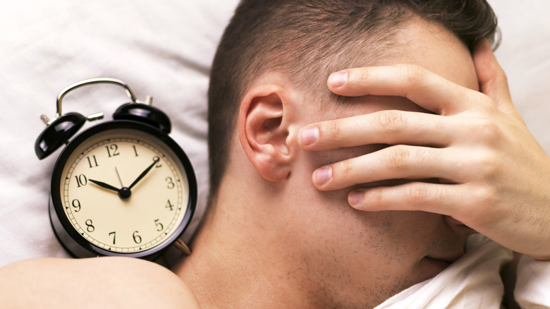 Young man covering his face with his hand sleeping next to an alarm clock