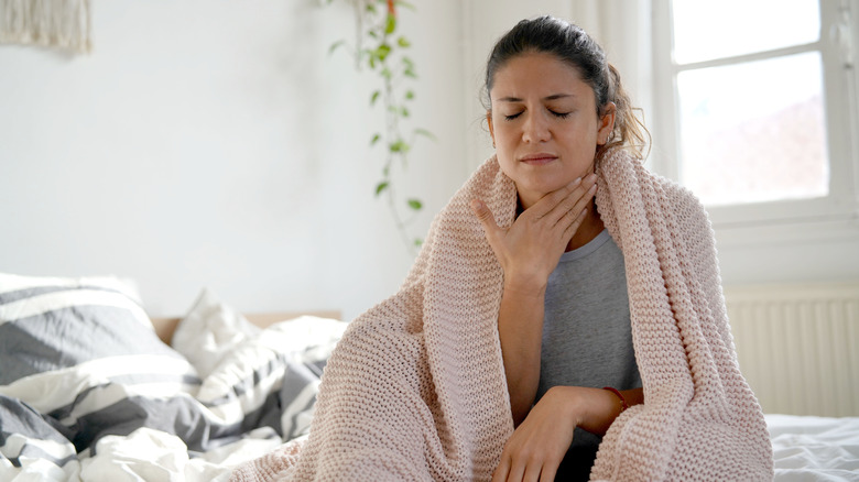 A woman sits on bed with a sore throat
