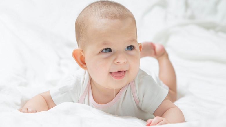 A baby in white clothes is lying on a white bed, smiling, drooling