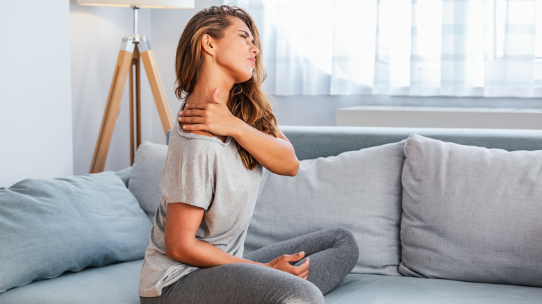 Woman in gray leggings and t-shirt sitting on sofa holding painful shoulder