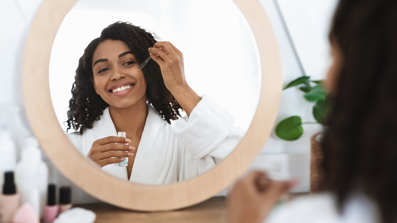 Woman smiles into mirror while applying serum to her face