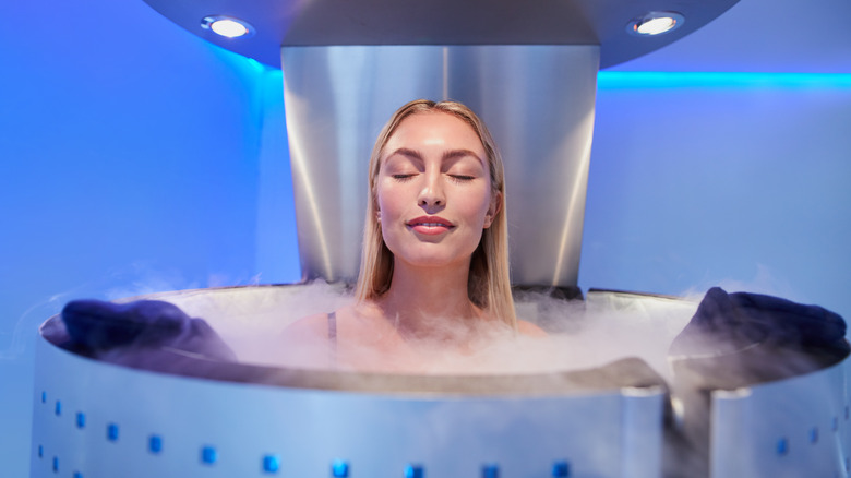 Woman with long hair and eyes closed, smiling in a metal cryotherapy chamber with liquid nitrogen gas coming out of the top. She is only visible from the shoulders up. 