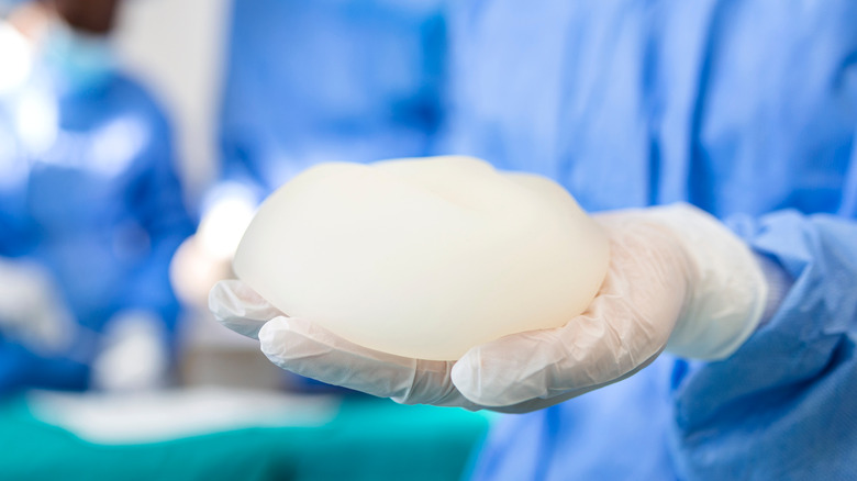Surgeon holding breast implant with 2 surgeons in blurred background performing surgery