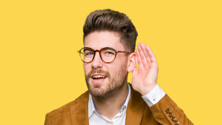 Young man wearing glasses smiling with hand behind ear trying to hear