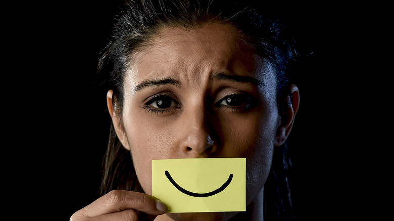 Distressed woman hides her anxiety behind a piece of paper with a smiley face held up to her mouth