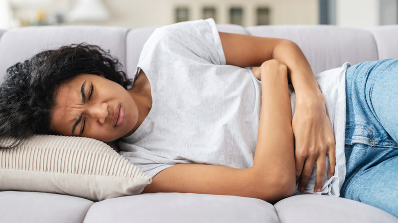 Young woman with dark hair and tan skin on a couch holding her stomach in pain