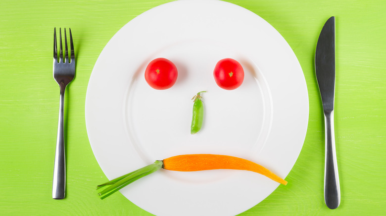 A sad face made of a carrot, a pea pod, and two tomatoes on a white plate