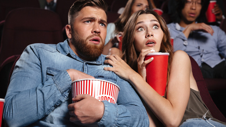 Couple eating popcorn at movie theater reacting to scary movie