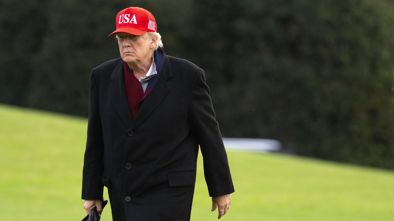 President Donald Trump in a red hat and black coat, walking on the grass with gloves in hand.