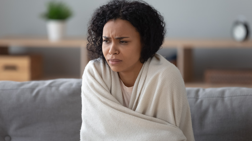 Person with curly brown hair sits on the couch wrapped in a white blanket looking uncomfortable 
