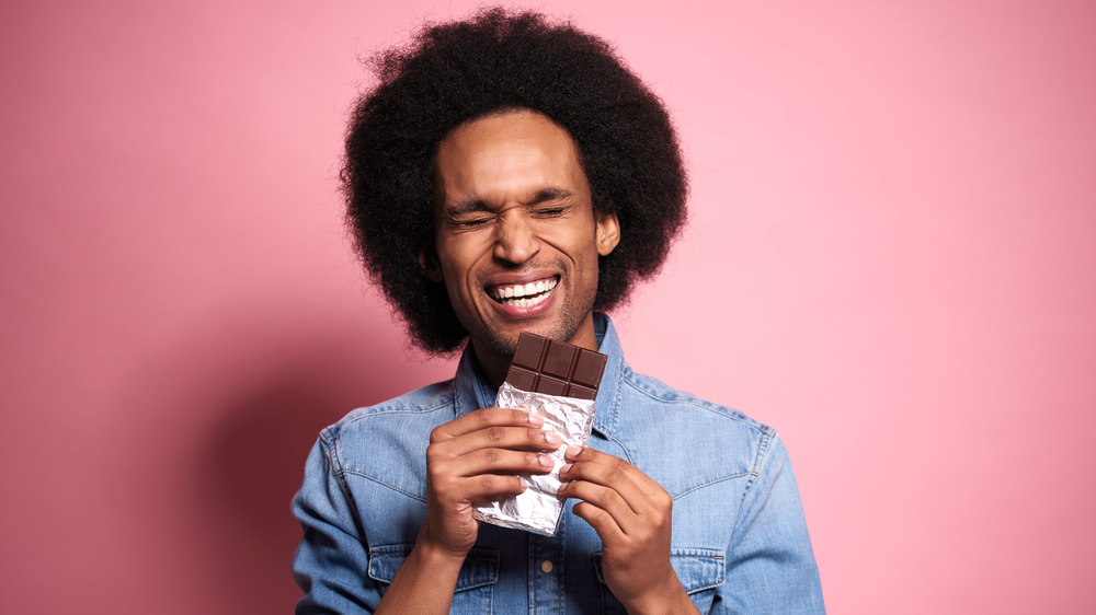 Person with dark hair and a blue shirt standing in front of a pink background holding a large chocolate bar and smiling with their eyes closed