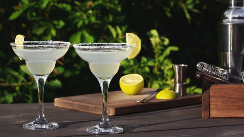 Two margaritas on an outdoor table next to cut limes and drink mixing equipment